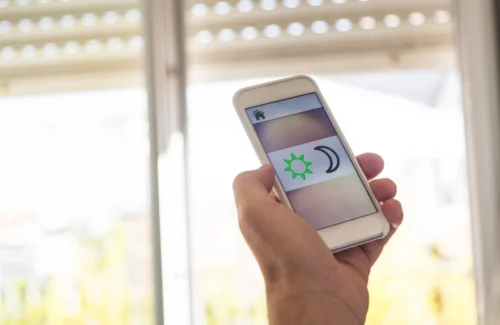 Hand mit Smartphone und App für Rollladensteuerung vor einem Fenster mit halb geschlossenem Rollladen