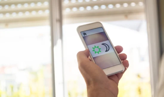 Hand mit Smartphone und App für Rollladensteuerung vor einem Fenster mit halb geschlossenem Rollladen